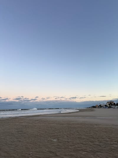 The image displays a coastal scene in Grottammare, Italy, facing the Adriatic Sea during twilight. The upper 60% of the frame is occupied by a clear sky, transitioning from a light blue at the zenith to a paler, slightly yellowish-pink hue at the horizon. A horizontal band of scattered stratocumulus clouds is present above the sea.

The middle ground features the sea, exhibiting moderate waves breaking onto the shore and against a series of dark, elongated rock groynes extending perpendicularly from the coastline. A prominent white-crested wave is breaking near the center-left, behind one of the groynes. The water appears a greenish-blue.

The foreground consists of a wide, unpopulated sandy beach, displaying a light brown coloration with subtle undulations and evidence of receding water lines. The beach curves gently from the center-left towards the right side of the frame. In the distant right, along the shoreline, a cluster of low-rise buildings with light-colored facades is visible. In front of these structures, indistinct white objects, consistent with beach umbrellas or tents, are discernible on the sand. Scattered palm trees are also visible near the distant buildings. No people, animals, or direct interactions are observed within the scene.