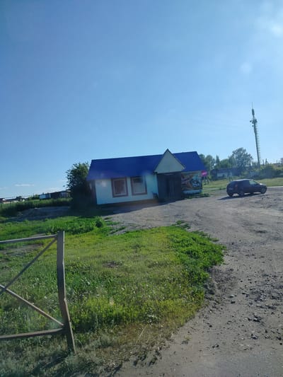 The image depicts a sunny day in a rural area, likely Степное Полеологово, Russia. The sky is a clear, vibrant blue, suggesting fair weather.
The main subject is a small, light-colored building with a dark blue roof. It appears to be a shop or small store, with "ПРОДУКТЫ" (PRODUKTY - meaning "Products" in Russian) visible above the entrance. There are also signs on the side of the building, one with Russian text that seems to include "Вкусный", which translates to "tasty". To the right of the building, a dark-colored hatchback car is parked on a gravelly dirt road.
In the background, a tall telecommunications tower stands prominently against the blue sky. Behind the shop, a dense line of trees and more buildings are visible, suggesting a small settlement or village. The foreground shows a grassy area with a metal fence, indicating a boundary or entrance to a property.
The overall scene conveys a sense of quiet, everyday life in a rural Russian setting on a bright, clear day. The presence of the shop and the car suggests ongoing activity, while the vast blue sky and open landscape evoke a feeling of peace and spaciousness. The time of day is clearly daytime due to the bright sunlight and clear skies.