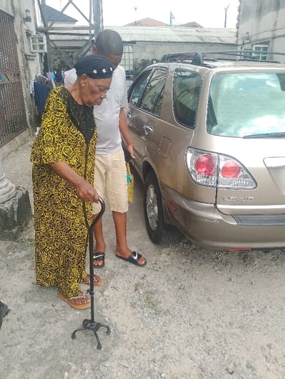 The image captures an outdoor scene in Mgbuoshimini, Nigeria, showing two individuals next to a parked gold Lexus SUV. An elderly woman, with dark skin and wearing a black and yellow patterned long dress, a dark head covering adorned with small white details, and light-colored flip-flops, stands in the foreground. She uses a black, four-footed cane held in her right hand for support and is looking downwards. A younger man, also with dark skin, stands slightly behind and to the right of the woman. He wears a light grey short-sleeved t-shirt, light brown shorts, and black Nike slide sandals, and is observing the woman. The rear driver's side of a gold Lexus SUV, equipped with roof rails, is visible to the right of the individuals. The vehicle bears "LEXUS" badging on its tailgate. The ground beneath them is unpaved, consisting of light-colored gravel and dirt. In the background to the left, a concrete pillar, a dark metal gate, clothes hanging on a line, and a white air conditioning unit on a light-colored building are discernible. Further back, corrugated metal roofs are visible under a bright sky. The interaction suggests the man is accompanying or assisting the woman.