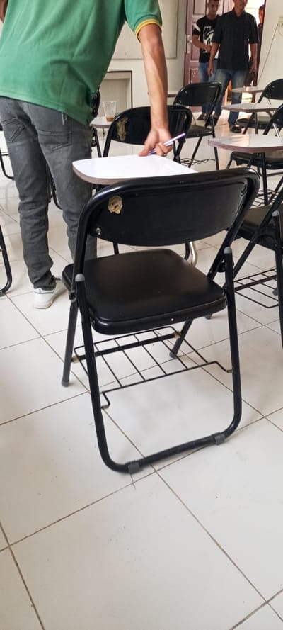 A man in a green shirt and gray jeans stands behind a black folding chair with a white writing surface. The chair has a black seat and a metal frame, with a wasp nest visible on the chair back. Behind the chair are other similar chairs and people. The floor is covered in white tiles. The person is holding a pen.