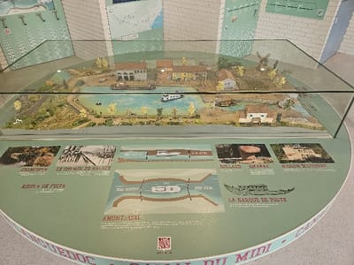 An indoor photograph captures a detailed miniature diorama of a canal section, presented under a transparent glass case on a circular, light green display table. This scene is located within a museum or interpretive center in Avignonet-Lauragais, France, indicated by contextual elements related to the Canal du Midi.
The diorama depicts a light blue waterway, bordered by green-brown land, small yellow-green trees, and narrow paths. Miniature structures include several multi-story buildings, one larger industrial-style building, and a windmill. A small bridge spans the canal. Multiple miniature boats are positioned within the canal; one larger barge is near the industrial building, and at least one smaller white boat is further along the canal. No active human figures are visible within the diorama.
The circular display table's surface features various informational graphics and images. These include photographs labeled 'L'ÉPANCHOIR' (spillway), 'LE CHEMIN DE HALAGE' (towpath), 'BOLLARD AMARRE' (mooring bollard), and 'MAISON ÉCLUSIÈRE' (lock house). A schematic diagram illustrates a canal lock system with a boat, marked 'AMONT / AVAL' (upstream/downstream). A line drawing of a boat is labeled 'LA BARQUE DE POSTE' (post boat). A QR code is present on the table, and the bottom edge features partial text reading 'CANAL DU MIDI'. The background consists of white walls with additional wall-mounted display graphics and maps. The area is illuminated by overhead interior lighting.