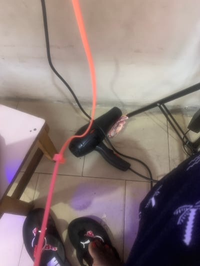 The image depicts an indoor scene, likely a room, on a tiled floor. A black hairdryer is visible, its cord extended and secured with a pink zip tie to a pink cable. The hairdryer is positioned near a black metal stand. A rolled-up wad of banknotes, appearing to be Euros, is tucked into the hairdryer's nozzle. To the left, a portion of a white table with wooden legs is present. In the foreground, a person's feet wearing black Puma flip-flops are visible. The overall lighting suggests ambient room light, with a purplish hue cast on the floor and table. The scene appears to be set in a residential or personal space within Rumuokoro, Nigeria.