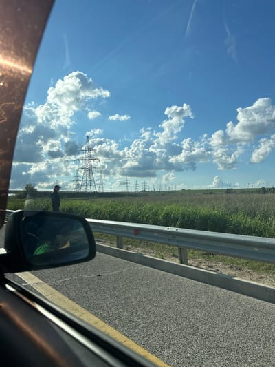 This media file depicts a rural landscape in Primorskii, Russia, viewed from a vehicle. The scene is dominated by a bright blue sky filled with dramatic, fluffy white cumulus clouds. In the distance, a field of tall, dry grass or crops stretches towards the horizon, punctuated by numerous tall power pylons, some with wires stretched between them. A metal guardrail runs along the right side of an asphalt road, which occupies the foreground. The sun is high in the sky, suggesting it is daytime, likely late morning or afternoon. The weather appears to be fair, with sunny conditions and scattered clouds.

A single person is visible in the mid-ground, standing in the field near the power pylons. They are wearing a hat and appear to be looking towards the power lines. The reflection in the car's side mirror shows a glimpse of the car's interior and possibly another passenger. The image has a slightly tilted perspective, as if captured from a moving vehicle. There is no visible text in the image. The overall mood is one of open space and rural tranquility under a vast sky.
