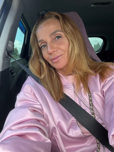 A woman with long, blonde hair and freckles is sitting in the passenger seat of a car. She is wearing a pink satin bomber jacket and a seatbelt is across her chest. The interior of the car is visible, including the door, window, and a purple fuzzy headrest. Outside the car, trees and a cloudy sky are visible. The image appears to be a selfie taken from inside the vehicle. There is no visible indication of the location being in Kerro, Russia.