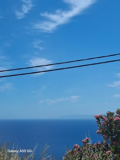 The image depicts a view of the sea and sky, likely taken in or near Gazi Municipal Unit, Greece. The sky is blue with wispy clouds. Two black power lines run diagonally across the left side of the frame. The sea is a darker shade of blue, meeting the sky at the horizon. In the upper right corner, there is foliage with pink flowers. The text "Galaxy A55 5G" is visible in the bottom right corner, indicating the device used to capture the photo. The image is rotated 90 degrees clockwise.