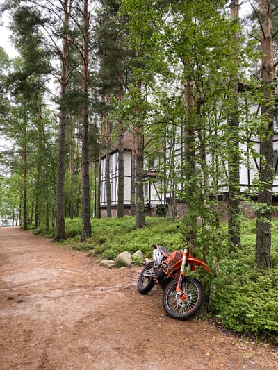 In Моторное, Russia, an orange and white dirt bike is parked on a dirt path next to a forest. The path is covered in pine needles and is lined with tall pine trees on the right. Behind the trees, a white building with dark trim and a stone foundation is visible. The dirt bike is leaning against a small tree and appears to be new. The surrounding vegetation is lush and green, with a mix of trees and bushes. The sky is overcast, suggesting a cloudy day.