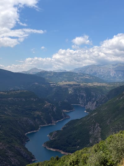 The image shows a panoramic view of a deep canyon with a winding, blue river flowing through it. The canyon walls are steep and covered with lush green trees. In the distance, several layers of mountains are visible, with the furthest ranges appearing hazy. The sky is blue with scattered white clouds. This scene is located near the city of Dubljevići, Montenegro, specifically depicting the Piva River Canyon. The image does not contain any people, objects, or actions.