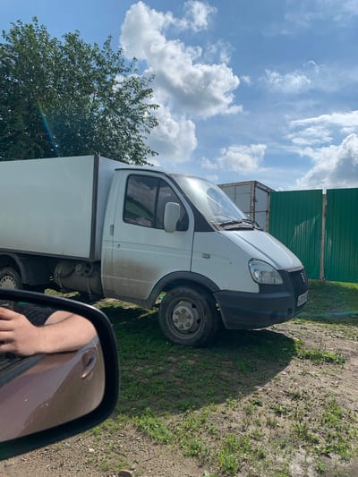 In Barsai, Russia, on a sunny day with scattered white clouds, a white delivery van with a cargo box is parked on a dirt and grass lot. The van is dirty, with mud splattered on its lower panels and wheels. A green corrugated metal fence stands to the right of the van, and a rusted shipping container is visible behind it. In the foreground, a side-view mirror of a car shows a person's arm and shoulder, indicating the photo was taken from inside another vehicle. A large tree with green leaves is on the left side of the frame, and a subtle rainbow arc is visible near the top left corner. The ground is a mix of gravel, dirt, and sparse green grass.