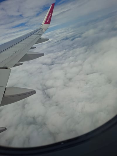 The image shows a view from an airplane window, looking out at the sky and the wing of the aircraft. The wing is predominantly white, with a pink section at the tip that features the text "Wizzair.com" in white. Several winglets are visible extending upwards from the main wing. The sky is filled with a dense layer of white, fluffy clouds. The scene suggests the aircraft is in flight, above the cloud layer. The lighting and the type of clouds indicate it could be daytime. There are no people visible in the image. The location context provided is Naantali, Finland.