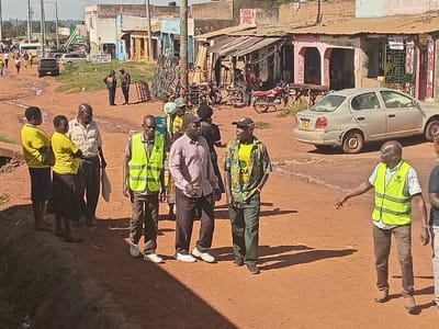The scene depicts a daytime view of an unpaved, reddish-brown dirt road in Shirungu-Mugai ward, Kenya. Approximately fifteen individuals are present. In the midground, a group of eight people stands or walks on the left side of the road, adjacent to a concrete culvert. This group includes three women uniformly dressed in yellow shirts and dark bottoms, an older man in a light-colored collared shirt, and two men, one wearing a lime-green high-visibility vest bearing an "LCCA" logo. Further right, two additional men are visible; one in a patterned short-sleeved shirt and a dark baseball cap, and another in a white shirt and a lime-green "LCCA" vest, extending his right arm in a gestural manner.
A light-colored Toyota Vitz sedan, displaying a Kenyan license plate, is parked on the right shoulder of the road, oriented away from the viewer. Behind this vehicle, a row of single-story commercial buildings with corrugated metal roofs lines the road. These structures feature open storefronts, with partially legible signage, including one indicating "SALON." A red motorcycle and various metal merchandise racks are positioned outside the shops. In the background, the dirt road extends, populated by additional pedestrians and vehicles, including a white van. Wooden utility poles with electrical wiring are interspersed along the roadside. The environment is consistent with a semi-urban commercial area under clear, sunny conditions, evidenced by distinct shadows.