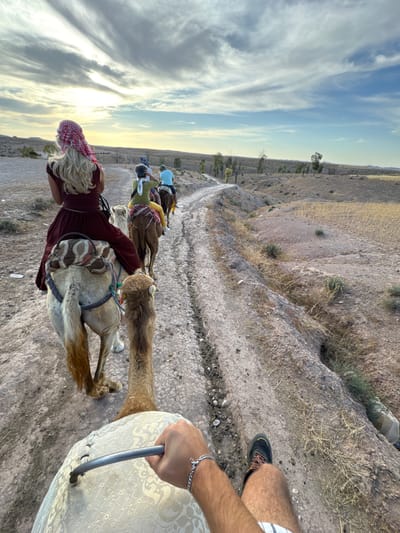 A person's perspective from riding a camel shows a group of people on camels moving along a dirt path. The person in the foreground is holding onto a metal bar attached to the camel's saddle. The person is wearing a bracelet and shorts. The camel in front is lighter colored, and the person riding it wears a red dress and a red and white patterned headscarf. The path leads through a dry, arid landscape with scattered trees and vegetation. In the distance, more camels and riders are visible. The sky is cloudy with patches of blue. The image appears to be taken in the late afternoon, with the sun casting a warm glow on the scene. The location is identified as Lalla Takarkoust, Morocco, which is known for its desert landscapes and camel trekking experiences.