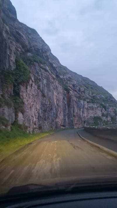 An overcast, dusk scene captures a view from a vehicle driving along a coastal road. The road curves to the right, bordered by a steep, rocky cliff face on the left. The cliff is streaked with darker and lighter tones, with patches of green vegetation clinging to its upper sections. The road surface appears wet, reflecting the headlights of the vehicle, which cast a bright, yellowish glow. The sky is a uniform grey. The image depicts the Great Orme's Head road in Llandudno, Wales.