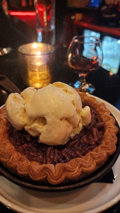An indoor scene depicts a pecan pie served in a small, dark cast-iron skillet, topped with five scoops of vanilla bean ice cream. The pie features a golden-brown, wavy, crimped crust and a dark, glossy pecan filling. The skillet rests on a white plate, positioned on a dark, flat surface. A dark, possibly black, napkin or cloth is visible beneath the skillet on the right side. In the blurred background to the left, a lit candle glows within a decorative, golden-toned votive holder, with a clear glass behind it. To the right, a stemmed snifter glass containing an amber liquid is visible. Further background elements are out of focus, showing indistinct shapes and ambient lighting, including a prominent horizontal red light bar and areas of blue and white illumination, consistent with a bar or restaurant environment. The overall lighting is dim. No individuals are present in the frame. The specific location within Jackson, United States of America, cannot be determined from the provided visual information.