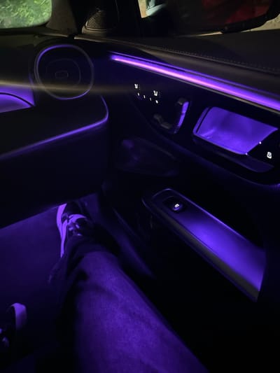 The image displays the interior of a passenger vehicle at night, illuminated predominantly by purple ambient LED lighting. The perspective is from within the vehicle, focused on the right-side door panel and a portion of the footwell, in Glendale, United States.
**Subjects/Objects:** The door panel features integrated light strips, emitting a violet glow along the upper trim, around a circular speaker grille, within the door handle recess, and illuminating the window control panel. A control cluster with three memory preset buttons (labeled 1, 2, M) and additional seat adjustment toggles are visible. Below these controls, a single window switch is present on a horizontal armrest segment, also bathed in purple light. The floor of the vehicle is similarly illuminated.
**People:** A human leg, clothed in blue denim jeans, extends into the lower half of the frame. The foot is clad in an athletic shoe, primarily black and white with white laces. The individual is situated inside the vehicle.
**Environment:** The car's interior lighting system casts a consistent purple hue across all visible surfaces, creating a distinct ambiance. Outside the upper right window, indistinct green foliage is faintly discernible, indicating an outdoor, likely parked, setting at night.