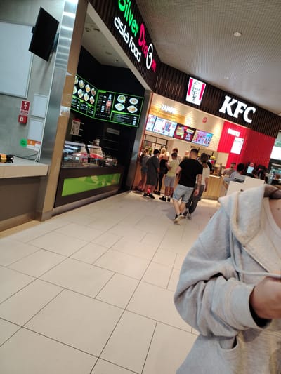 The image shows a food court area, likely within a shopping mall in Wołomin, Poland. To the left, there is a food stall with a digital menu displaying Asian food options. Further to the right, a KFC restaurant is visible, with its distinctive logo and red branding. A group of people are standing in line, presumably waiting to order food from either establishment. The floor is tiled with large, light-colored square tiles. The lighting appears to be artificial, typical of an indoor shopping environment. The perspective suggests the photo was taken from a slightly elevated position, possibly by someone standing in the food court.