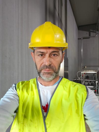 A man wearing a yellow hard hat and a neon yellow safety vest stands in front of a concrete wall, holding a camera for a selfie. He has a beard and is wearing a white sweatshirt with a red maple leaf emblem. Behind him, industrial equipment, including pipes and a metal rack, are visible. The scene appears to be inside a building. The location is identified as Warcoing, Belgium.