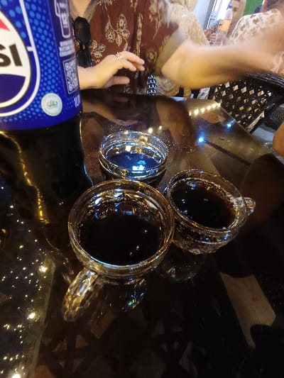 The media file captures a close-up, overhead view of a table with drinks and a person's hand. Three glass cups, filled with a dark liquid, are arranged on a reflective, dark surface, likely a table. The cups appear to be holding a cola-like beverage, as indicated by the dark color and visible bubbles. One cup is larger and has a handle, while the other two are smaller and handleless.
In the foreground, a portion of a blue and white bottle is visible, suggesting it might be the source of the drinks. The bottle has text and a logo on it, and a circular green emblem with an Arabic inscription is also present. The surface of the table reflects lights, creating a bokeh effect that adds to the ambiance.
A person's hand with a ring on one finger is visible reaching towards the table, and another hand is partially visible in the upper left corner of the image. The person's attire, a patterned fabric, is also visible. Sunglasses are placed on the table near the bottle.
The scene appears to be set indoors, possibly in a restaurant or cafe, given the lighting and the presence of drinks. The lighting suggests it could be evening or nighttime, with artificial lights creating reflections. There are no clear indications of weather conditions. The overall mood seems relaxed, possibly a social gathering or a break. The location context of Nematjan, Uzbekistan, is provided separately and is not directly inferable from the visual cues in the image.