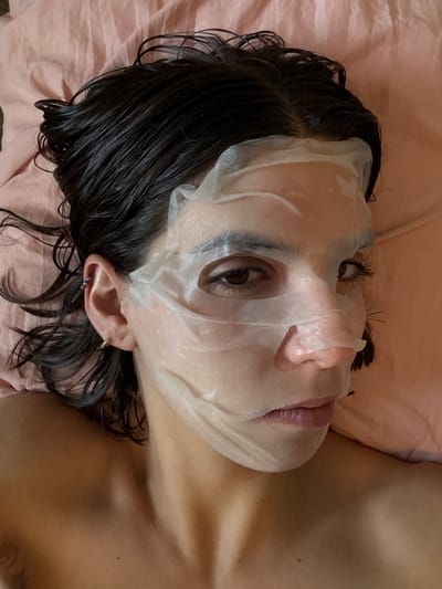 A woman lies on a pink pillow in Southlake, Texas, USA, wearing a white sheet mask. Her wet, dark hair is scattered on the pillow around her head. She has an earring in her left ear, and parts of her upper chest and shoulder are visible. The focus is on her face, which is partially covered by the mask.
