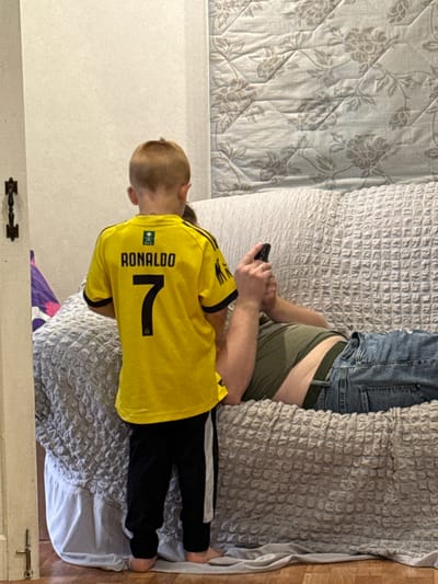 A young boy wearing a yellow Ronaldo jersey and black pants stands facing away from the camera, looking at a person lying on a couch. The person on the couch is wearing a green t-shirt and blue jeans and is holding a smartphone. The couch is covered with a grey textured blanket. The background consists of a wall and a floral-patterned quilt. The scene appears to be indoors, possibly a living room.
