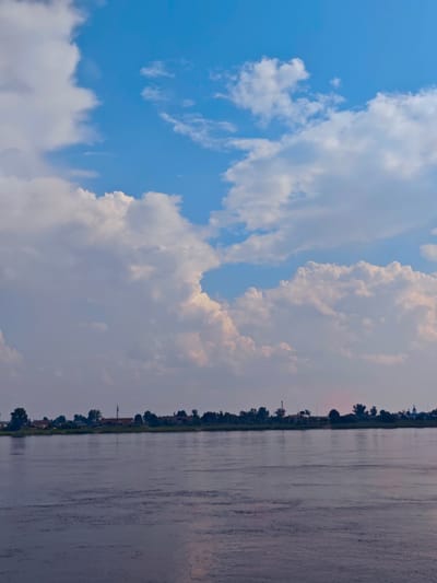 The image depicts a wide, calm river with a gently rippling surface. The water reflects the soft hues of the sky and the distant shoreline. On the far bank, a line of trees and houses is visible, suggesting a rural or semi-rural settlement. The sky above is a vibrant blue, dotted with large, fluffy white clouds that cast soft shadows on the landscape below. The overall impression is one of tranquility and natural beauty.
This scene is not located in Yeniseysk, Russia. The architecture and vegetation do not match typical characteristics of that region.