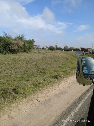 A rural landscape under a partly cloudy sky. A grassy hillside meets a dirt shoulder beside a paved road. A sign reading "KAJIADO" indicates the location. In the distance, utility poles and structures dot the horizon. The scene is captured from inside a vehicle, evidenced by the side mirror and interior details visible in the lower right corner. The date and time stamp "2025/11/22 08:16" appear at the bottom.