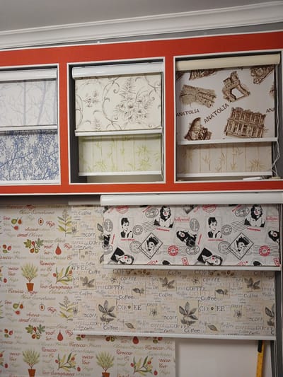 The image shows a display of roller blinds with various patterns. The blinds are arranged in sections, with three visible on the top row and one on the bottom row. The top row features blinds with a gray tree silhouette pattern, a floral pattern, and an Anatolian architectural pattern. The bottom row displays blinds with a pattern of fruits (figs and olives), coffee cups, and Hollywood icons like Marilyn Monroe and Audrey Hepburn. The blinds are mounted on a wall with a red border. There are no people or specific actions depicted. The environment appears to be a commercial setting, possibly a store or showroom, where window treatments are displayed. There is no information that can be used to determine the specific location within the City of Türkmenabat, Turkmenistan.