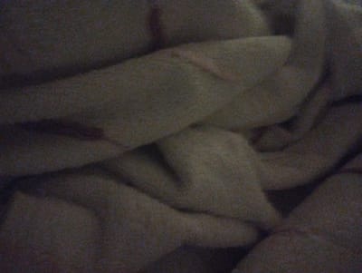The image is a close-up, out-of-focus shot of a crumpled, light-colored fabric, likely a blanket or bedsheet. The texture of the fabric is visible, with a grainy appearance due to low lighting and image quality. There are folds and shadows that create a sense of depth and volume, suggesting the fabric is draped or bunched up. No specific location in Sumgayit, Azerbaijan, can be identified from this image, as it lacks any recognizable landmarks or contextual elements. The scene is devoid of any people, objects, actions, or interactions.