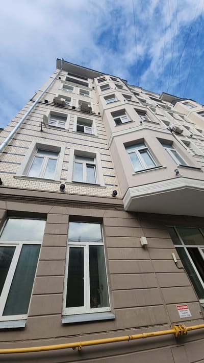 This media file captures an upward view of an apartment building in Uzlovaya, Russia. The building features a light-colored facade with several rows of windows. The architecture is modern, with some decorative elements around the windows and balconies. A prominent yellow gas pipe runs vertically along the right side of the building.

On the brown-paneled section of the building, to the right of the windows, there is a red and white warning sign. The text on the sign is in Russian and reads "ОСТОРОЖНО ВОЗМОЖНО ПАДЕНИЕ СНЕГА И НАЛЕДИ", which translates to "CAUTION, SNOW AND ICE MAY FALL".

The sky above is blue with scattered white clouds, suggesting a daytime scene with fair weather. The perspective, looking upwards, emphasizes the height of the building and the vastness of the sky. There are no people visible in the frame, and no specific activity is taking place. The overall impression is one of an ordinary urban environment in a Russian city.