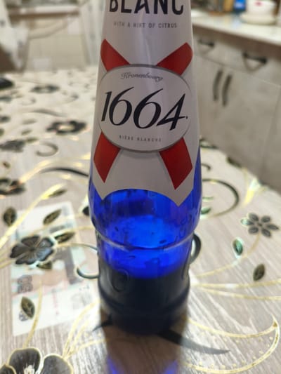 The media file shows a close-up of a Kronenbourg 1664 beer bottle with a blue liquid inside, resting on a patterned tablecloth. The bottle has a white label with red accents and text. Visible text on the label includes "BLANC", "WITH A HINT OF CITRUS", "Kronenbourg", "1664", and "BIERE BLANCHE". In the background, blurred kitchen cabinets are visible, with the overall lighting suggesting an indoor setting, possibly a home or restaurant. To the left of the bottle, a portion of Russian currency, specifically a 50-ruble banknote, is visible on the tablecloth. The scene is static, with no active event taking place. The focus is on the product and its presentation. The mention of Luxembourg and Kyrgyzstan in the prompt does not seem to be directly supported by visual cues within the image itself, other than the potential origin of the banknote.