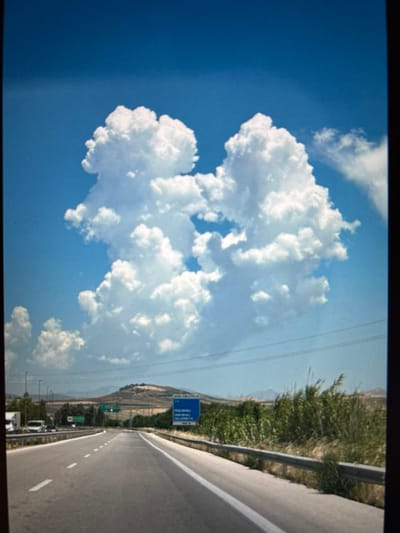The image shows a bright blue sky filled with large, fluffy white cumulus clouds. The camera is positioned at an angle, creating a sense of perspective. To the right, a paved road with white dashed lines curves away into the distance. A metal guardrail runs alongside the road. On the left side of the road, there are green bushes and trees, with a hillside in the background. A blue road sign with white lettering is visible on a pole. The overall scene suggests a sunny day, possibly in the late morning or early afternoon, given the brightness and the distinct shadows. The setting appears to be a rural or semi-rural area, likely a highway or main road. There are no people visible in the image. The image's orientation is on its side.