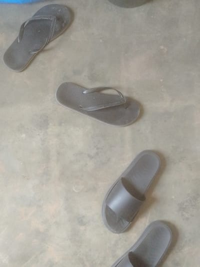 In Majemu, Nigeria, on a dull, concrete floor, four dark-colored flip-flops and slides are scattered. Two flip-flops are positioned near the top left of the image. One is open, with its strap extended upwards, and the other is closed, showing its sole. Below these, towards the center, another pair of flip-flops lies open. The lower right portion of the image features two slides, one directly above the other, both facing towards the bottom. The floor exhibits a mottled texture with variations in shade, suggesting wear and tear. There are no people or animals present in the scene.