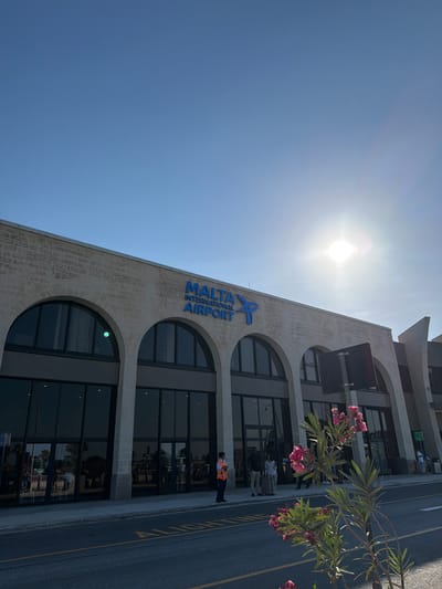 The image depicts the exterior of Malta International Airport on a bright, sunny day. The sky is a clear, vivid blue, with the sun prominently visible, suggesting it is likely midday. The building's facade is made of light-colored stone with large, arched windows. The words "MALTA INTERNATIONAL AIRPORT" are displayed in large, blue, stylized lettering on the side of the building.
In front of the building, a paved road runs alongside the sidewalk. Several people are visible near the entrance of the airport, some walking and others standing. One individual, wearing a bright orange safety vest, is standing on the sidewalk near the road, possibly a worker or security personnel. The reflection in the large windows shows more people inside the building. Lush green foliage with clusters of pink flowers is present in the foreground, adding a touch of color and nature to the scene. The overall impression is one of a modern, functional airport on a pleasant day.