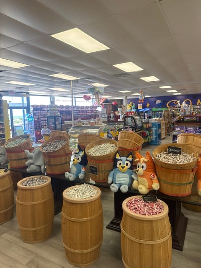 This is a medium shot of the interior of a candy store in Ocean City, Maryland, United States of America. The store is well-lit with fluorescent ceiling lights. Several large wooden barrels filled with different types of candy are prominent in the foreground and midground. The barrels are stacked and arranged on tables.
In the foreground, there are two wooden barrels. The left barrel is filled with small, individually wrapped candies, likely mints or sour candies. The right barrel is also filled with similar individually wrapped candies. Behind these, and to the left, is another large wooden barrel containing a grey, stuffed elephant toy.
In the midground, there are more wooden barrels. A barrel on the left is filled with grey and white striped candies. Next to it, another barrel contains a blue stuffed dog toy, possibly from the show Bluey. To the right of that, a barrel holds an orange stuffed dog toy, also likely from Bluey. Another barrel in the midground, to the right, is filled with coconut-flavored candies, as indicated by a sign that reads "Coconut". A sign above another barrel reads "Apple".
In the background, shelves are stocked with various candies, including M&M's, Milka chocolate bars, and other boxed candies. A glass display case is visible, showing what appear to be pastries or other confectionery items. A refrigerated beverage machine is also present. In the far background, through large windows, a glimpse of the outdoors is visible, suggesting a coastal location. The overall impression is a vibrant and abundant candy store.