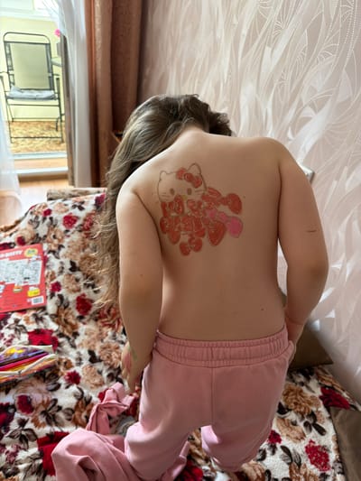 A young person is standing with their back to the camera, displaying a temporary tattoo of Hello Kitty on their upper back. The tattoo is outlined in a reddish-brown color and filled with pink and red hues, with the character's signature bow visible at the top. The person is wearing pink sweatpants. The setting appears to be indoors, with a floral patterned blanket covering a piece of furniture. A coloring book and a pencil case are visible on the blanket to the left. In the background, there is a window with a view of an outdoor area and curtains. The wall behind the person is decorated with a textured, light-colored wallpaper. There is no indication of the location being Valozhyn, Belarus, or any other specific city.