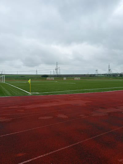 A wide-angle exterior view depicts an athletic complex in Suvorovo, Bulgaria, under an overcast sky. In the foreground, a wet, reddish-brown synthetic running track with white lane markings occupies the lower half of the frame. Beyond the track, a vibrant green artificial turf football pitch is visible, featuring white boundary lines, a white goalpost on the left side, and a yellow corner flag near the track. On the far side of the football pitch, approximately five white, covered dugouts or player benches are evenly spaced. The background consists of a flat, vegetated landscape, above which several large, metallic electricity pylons supporting overhead power lines are prominently visible, extending horizontally across the horizon. No individuals are present in the scene.