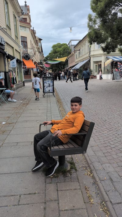 The image shows a young boy with dark hair and an orange hoodie sitting on a dark bench. He is looking at the camera with a slight smile. He is wearing dark jeans and black and white sneakers. The bench is on a paved sidewalk. In the background, there are shops with awnings, people walking, and trees. Some colorful banners are hanging across the street. The sky is cloudy. The scene appears to be in a town or city, likely during the daytime. Visible text includes what appears to be shop signs and a poster in a display stand. One sign reads "JERSEYLAGET" and another has a logo with "STORSJÖN". The overall impression is a casual street scene with a focus on the boy on the bench.