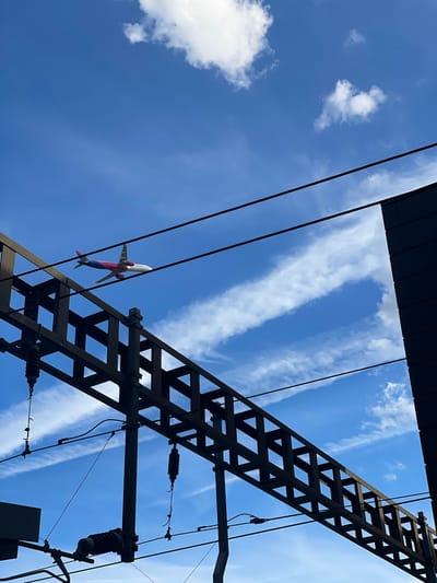 An airplane with pink and blue markings flies across a bright blue sky, partially obscured by thin, wispy clouds. In the foreground, the dark silhouette of electrical infrastructure, likely related to a railway line, dominates the lower portion of the frame. This structure consists of dark metal beams and wires, with some insulators visible. The scene suggests an outdoor, possibly urban, environment, with the sky indicating daytime. This specific scene is located near Luton Airport in Luton, United Kingdom, where air traffic is common.
