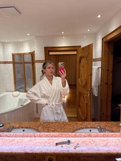 A woman in a white bathrobe is taking a selfie in a bathroom with a marble countertop. The bathroom has a jacuzzi tub with glass doors and wooden trim. The mirror reflects a wooden door leading to a sauna, with towels hung on a rack. Jewelry, including a watch and necklace, are placed on the countertop next to the sink.