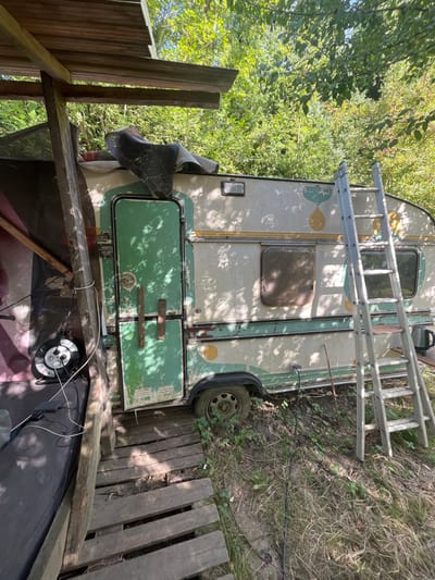The scene depicts a weathered, vintage-style travel trailer or caravan, predominantly off-white with peeling green and yellow/orange accents, situated outdoors within a densely vegetated area consistent with a rural or semi-wild environment in Sint-Pieters-Leeuw, Belgium. The precise location within the municipality cannot be determined from the image.
The caravan features a distressed green door with two vertical handles and a single rectangular window on its side. A black tarp is draped over the front portion of its roof. One wheel is partially sunken into the ground, indicating it has been stationary for an extended period.
To the left of the caravan, a rudimentary lean-to structure constructed from rough wooden posts and beams provides a partial overhead cover, supporting a corrugated roof section. Beneath this structure, a black mattress or large cushion is visible on the ground. Adjacent to the mattress, a white power extension cord reel with multiple outlets is present, connected to a black electronic device via several cables.
To the right of the caravan, an aluminum step ladder leans against its side, extending vertically beyond the roofline. A dark-colored kettle rests on a small, dark surface at the base of the ladder.
The ground immediately surrounding the caravan is uneven, composed of dirt, dry grass, and various green weeds. A makeshift pathway constructed from wooden pallets or planks leads to the caravan's entrance. The background consists of dense, lush green trees and foliage, illuminated by direct sunlight, which casts dappled shadows across the scene. No individuals are present, nor are any specific actions or interactions depicted.