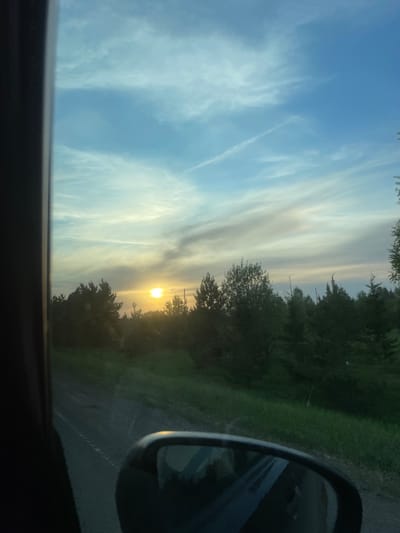 The image appears to be taken from inside a moving vehicle. The view is toward the horizon with the sun setting through trees. The sky has streaky clouds. A side mirror of the vehicle is visible on the right side of the image. No people are visible. There is no information to confirm the location as Kuregovo, Russia.