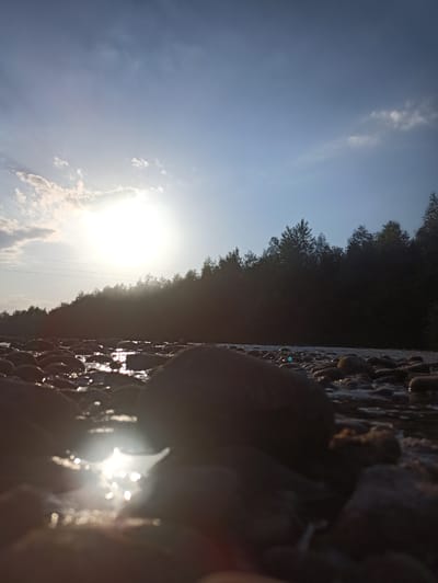 A low-angle shot depicts a sunlit riverbed in Kniazhdvir, Ukraine. The foreground is dominated by rounded, dark-colored river stones, some partially submerged in shallow water. Sunlight glints off the water's surface and the wet stones, creating bright reflections. In the middle ground, a dense line of dark green trees forms a silhouette against the bright sky. Overhead, the sky is a gradient of pale blue, with scattered, wispy white clouds on the left side near the sun. The sun itself is a bright, hazy orb positioned in the upper left quadrant of the image, casting lens flares and contributing to the overall bright and somewhat washed-out appearance of the sky. Subtle power lines can be seen stretching horizontally across the middle ground, just above the treeline. The scene is devoid of any human presence or activity.