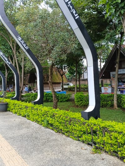 A serene park scene features curved, black and white arches with Arabic script amidst lush greenery. A neatly manicured hedge with a low, dark-colored fence lines the foreground, bordering a textured, gray walkway with yellow tactile paving. Beyond the arches, a traditional Javanese-style structure with a steeply pitched roof stands amongst trees. People are seated in the background. A small building to the right sells goods with signage visible.