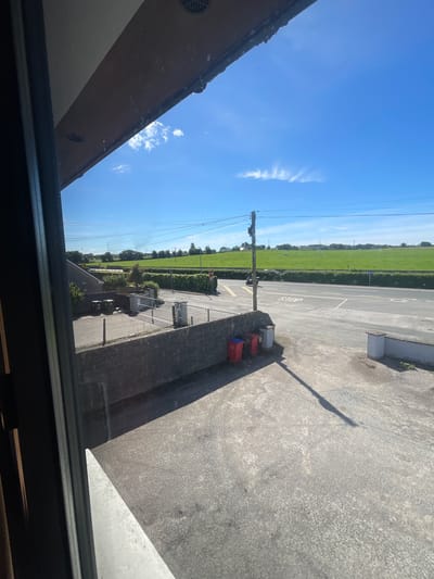 The image captures an outdoor scene on a sunny day, likely in Ireland, given the presence of a "STOP" sign and typical rural landscape. In the foreground, a rough concrete surface is visible, with a distinct shadow cast by an unseen object, possibly a utility pole. To the left, a dark, window-like frame suggests the vantage point is from indoors, looking out.
A low, grey stone wall runs horizontally across the middle ground, bordering a paved area. On this paved area, two large, green wheelie bins and a smaller red one are situated. Behind the wall, a road is visible with a "STOP" sign on the right side, indicating a junction. A grey sedan is parked or driving on the road.
Beyond the road, a vibrant green field stretches into the distance, bordered by neatly trimmed hedges. In the background, more fields and trees are scattered across a gently rolling landscape. The sky is a clear, bright blue with a few wispy white clouds and power lines crisscrossing the scene, typical of the Irish countryside. The overall impression is one of a peaceful, rural setting on a clear, pleasant day.