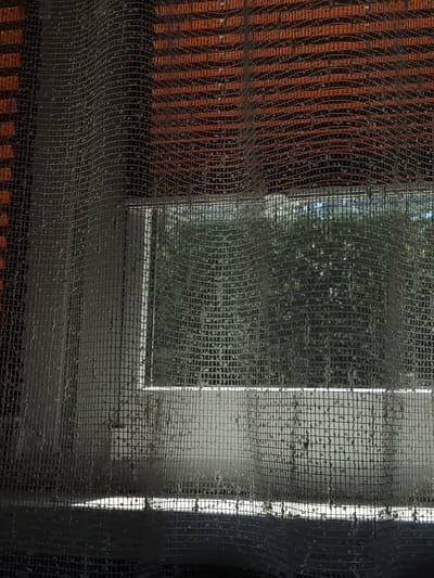 The image shows a window covered by a mesh screen with water droplets. Behind the screen, blinds with horizontal orange and dark brown stripes are visible. A white window frame is also present, and beyond that, green foliage can be seen, suggesting an outdoor environment. The lighting indicates it might be daytime, possibly overcast or with the sun filtered. The scene appears to be an interior view looking outwards, with the focus on the textured surface of the screen and the subtle details of the water on it. The location in Sosnowiec, Poland, is not directly evident from the visual content of the image itself, as there are no specific landmarks or identifying features that pinpoint this exact city.