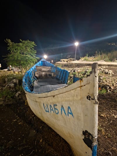 The image shows a weathered, white and blue fishing boat named "ШАБЛА" in the city of Tyulenovo, Bulgaria, at night. The boat is pulled ashore and rests on dark soil and rocks. Its blue interior is visible, with wooden benches and seats. The boat's hull shows signs of age and wear, with chipped paint and repaired sections. In the background, two streetlights illuminate the scene, casting long beams of light into the dark sky. Lush green vegetation, including a tree on the left, surrounds the boat and stretches up a gentle slope. The ground is covered with dirt, stones, and sparse grass.