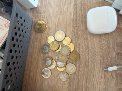 The scene, set on a light brown wooden surface, depicts an assortment of objects. To the left, a gray, perforated plastic storage container is partially visible, containing indistinct items, including a white and red paper fragment. Scattered across the mid-ground and right are approximately 22-25 Euro coins of various denominations, including 1 Euro, 2 Euro, and various cent coins, some bearing national designs like an eagle. In the upper right quadrant, a white rectangular electronic power adapter is positioned. Adjacent to the adapter and slightly below, a white, rounded rectangular object, consistent with an AirPods charging case, rests on the surface, displaying a subtle vertical seam. A white USB-C charging cable, with its connector end visible, extends from the bottom right towards the center of the frame. No individuals are present, and no active actions or explicit interactions are depicted. The implied location is Kermen, Bulgaria.