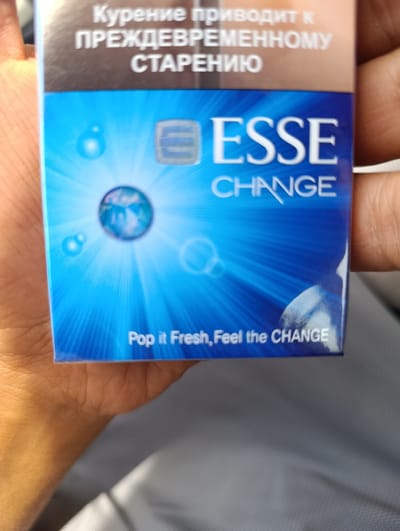 The image shows a close-up of a cigarette pack held in a person's hand. The pack is blue with white text. The text at the top of the pack is in Russian and reads "Курение приводит к преждевременному старению" which translates to "Smoking leads to premature aging". Below this, in large white letters, is the brand name "ESSE" and below that "CHANGE". Towards the bottom of the pack, in smaller white letters, it says "Pop it Fresh, Feel the CHANGE". The background is out of focus and appears to be a light gray fabric. There is no discernible location in the city of Khuroson, Tajikistan.