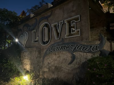 The image shows a large, illuminated sign in the shape of the word "LOVE" mounted on a textured stone wall. Above and to the left of the "LOVE" sign, the word "Bukit" is written in a stylized script. Below the "LOVE" sign, the words "Karimun Jawa" are written in a similar script. The letters of "LOVE" are three-dimensional and appear to be made of white material with dark outlines, lit from the front, casting subtle shadows on the wall. The sign is set against a backdrop of dark blue sky and lush green foliage. The scene is lit by artificial lights, including a bright spotlight near the bottom left illuminating plants and a portion of the wall. The overall impression is that of a scenic viewpoint or a tourist attraction in the evening. The location is identified as Karimunjawa, Indonesia. The text visible includes "Bukit", "LOVE", and "Karimun Jawa". There are no people visible in the image. The time of day is clearly night.