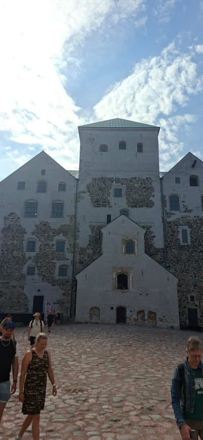 The image shows a group of people walking on a cobblestone courtyard in front of a historic stone castle. The castle has a prominent tower and multiple stone walls with visible signs of age, including weathered stones and plaster. Several windows of various sizes are visible on the castle walls. The sky above is partly cloudy with a mix of blue sky and white clouds. In the foreground, several individuals are walking towards the viewer and away from the castle. One woman is wearing a patterned dress, and a man next to her is wearing a t-shirt and shorts. Further back, more people are walking towards an entrance. The castle is located in Turku, Finland, and is likely the Turku Castle, a medieval building with a long history.