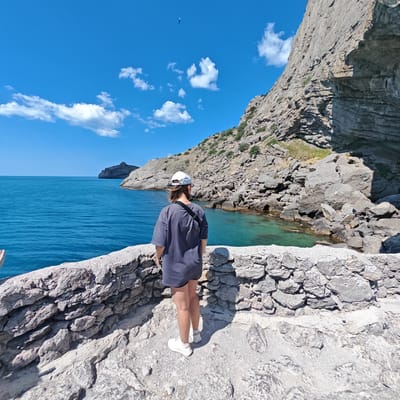 A person wearing a grey t-shirt, white cap, and white sneakers stands on a stone pathway, looking out at a clear blue sea and a rugged, rocky coastline. The person is seen from behind, with their back to the camera, and has a black bag slung across their shoulder. The pathway is made of rough stones and appears to be part of a scenic overlook or trail.
The sea is a vibrant turquoise, with some areas of deeper blue, and small waves gently lap against the shore. In the distance, a rocky island or promontory juts out from the water. The coastline is characterized by steep, grey cliffs covered in sparse vegetation. The sky is a bright, clear blue with a few scattered white clouds, suggesting a sunny day. A small object, possibly a kite or paraglider, is visible in the far distance against the sky. The overall scene evokes a sense of peaceful exploration and appreciation of natural beauty.
The time of day appears to be midday, given the bright sunlight and the shadows cast on the ground. The weather is clearly fair and warm. The location cues, such as the distinctive rocky cliffs and the vibrant blue sea, are consistent with a coastal Mediterranean or Black Sea environment. No visible text is present in the image.